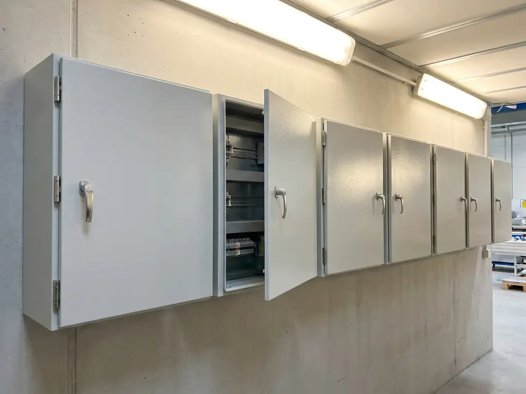 Wandmontierte Stahlschaltschränke in Hellgrau an Fabrikwand, ein Gehäuse leicht geöffnet mit sichtbaren Montageschienen.